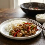 Plate of glossy black pepper chicken with onion and bell peppers over white rice with chopsticks